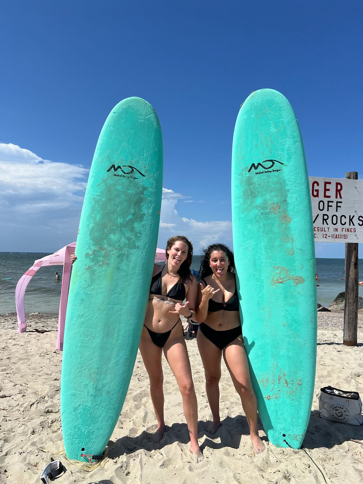 Two friends in black bikinis posing with tall turquoise surfboards on a sunny sandy beach, ocean and blue sky in the background, flashing shaka signs.