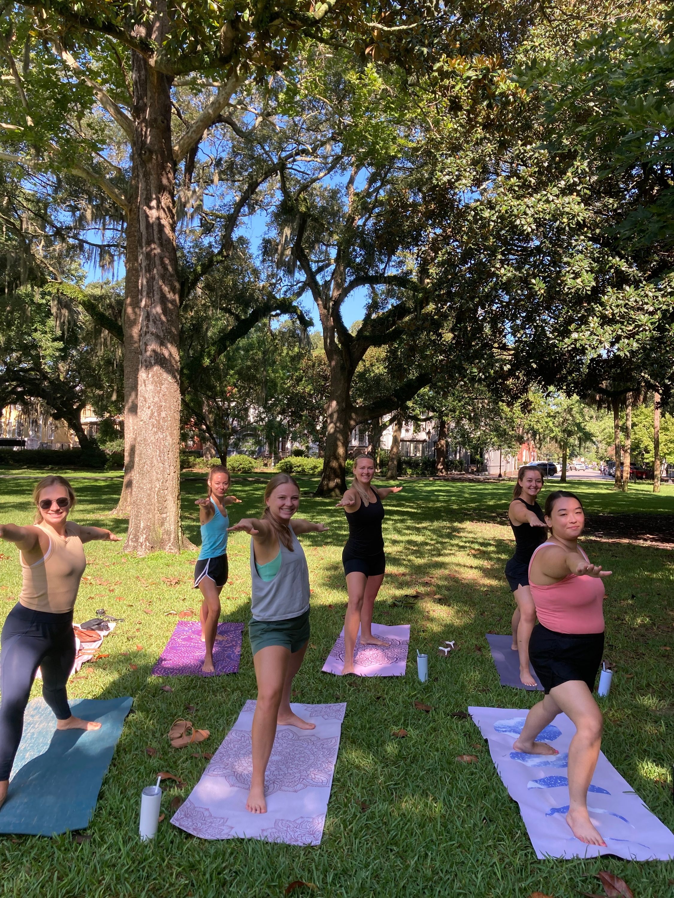 Six people practicing Warrior II yoga on colorful mats in a sunlit grassy park beneath sprawling oak trees draped with Spanish moss.