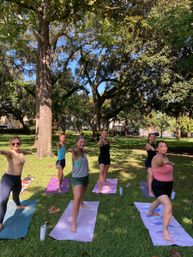 Six people practicing Warrior II yoga on colorful mats in a sunlit grassy park beneath sprawling oak trees draped with Spanish moss.