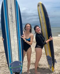 Two friends in swimsuits smiling and holding blue-and-white and yellow-and-black surfboards on a sandy beach with ocean waves and a cloudy sky