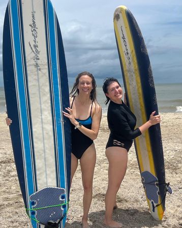 Two friends in swimsuits smiling and holding blue-and-white and yellow-and-black surfboards on a sandy beach with ocean waves and a cloudy sky