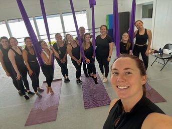 Instructor selfie with a cheerful group of women in an aerial yoga class at a bright studio, purple silks hanging above patterned yoga mats.