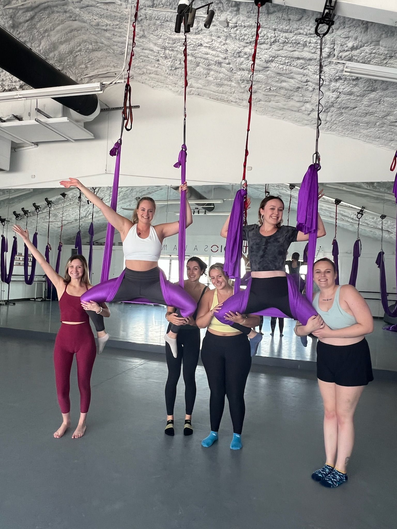 Group aerial yoga class in a bright studio — six women smiling as two are suspended in purple silk hammocks doing straddle poses while friends support them in front of a mirrored wall.