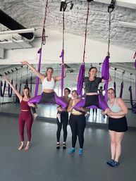 Group aerial yoga class in a bright studio — six women smiling as two are suspended in purple silk hammocks doing straddle poses while friends support them in front of a mirrored wall.