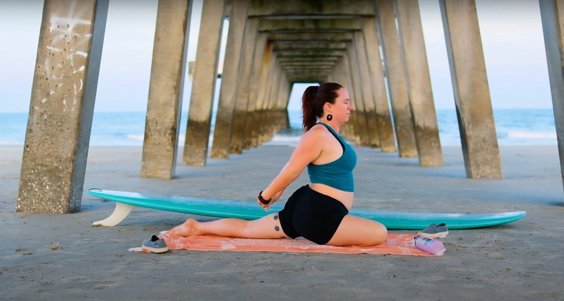 Person practicing beach yoga in a seated stretch on a towel next to a turquoise surfboard under a coastal pier on a sandy ocean beach