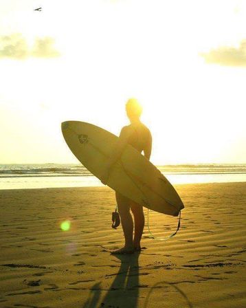 Silhouette of a surfer holding a surfboard on a sandy beach at golden sunset, ocean waves and long shadow in warm coastal light