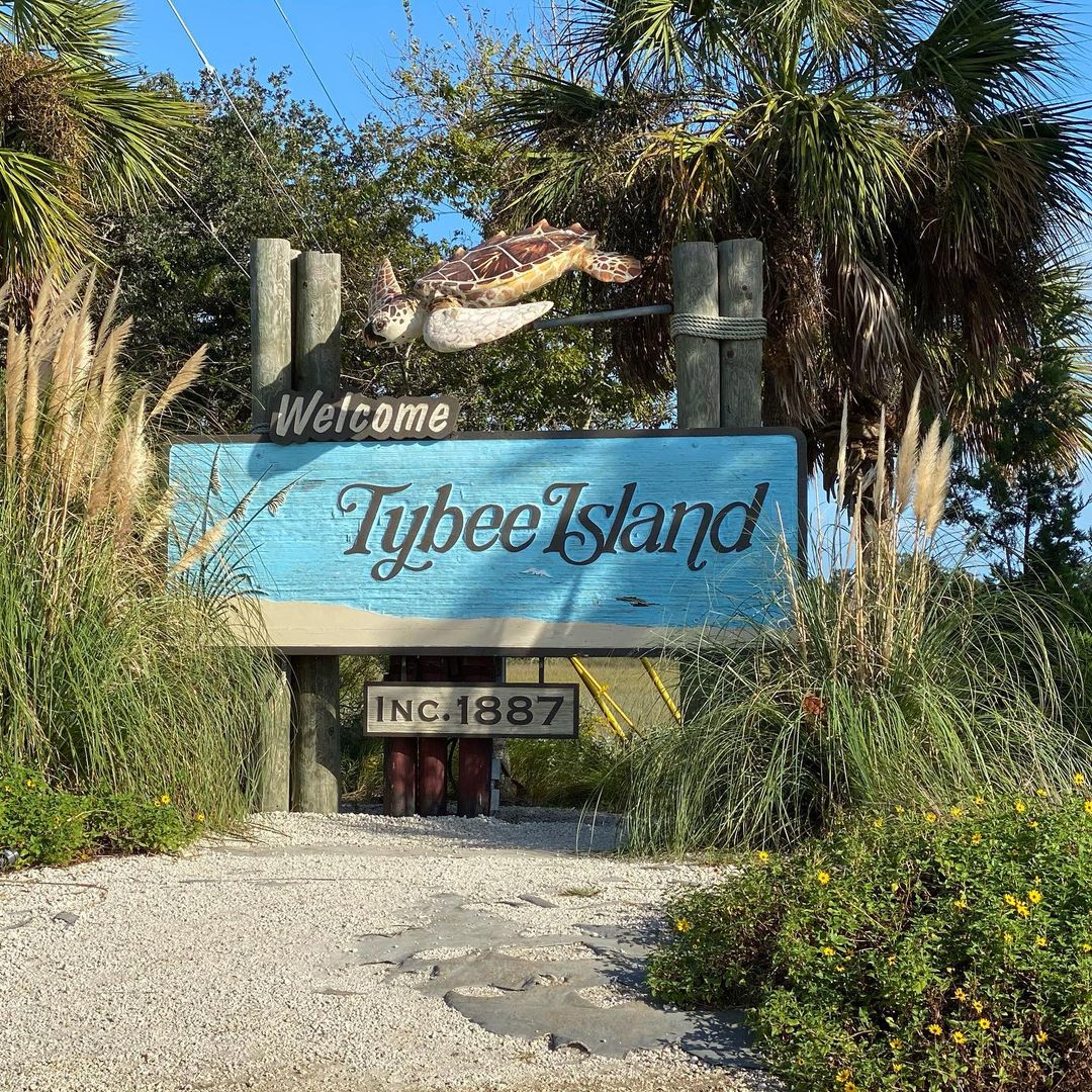 Welcome sign for Tybee Island — blue wooden board topped by a carved sea turtle, flanked by palm trees, coastal grasses and a sandy path; established 1887.