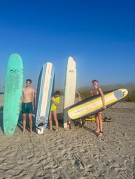 Four young people holding colorful surfboards on a sunny sandy beach with dune grass and a clear blue sky, ready to surf