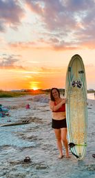 Smiling surfer in a red top holding a longboard on a sandy beach at a pink-orange coastal sunset with low dunes on the horizon.