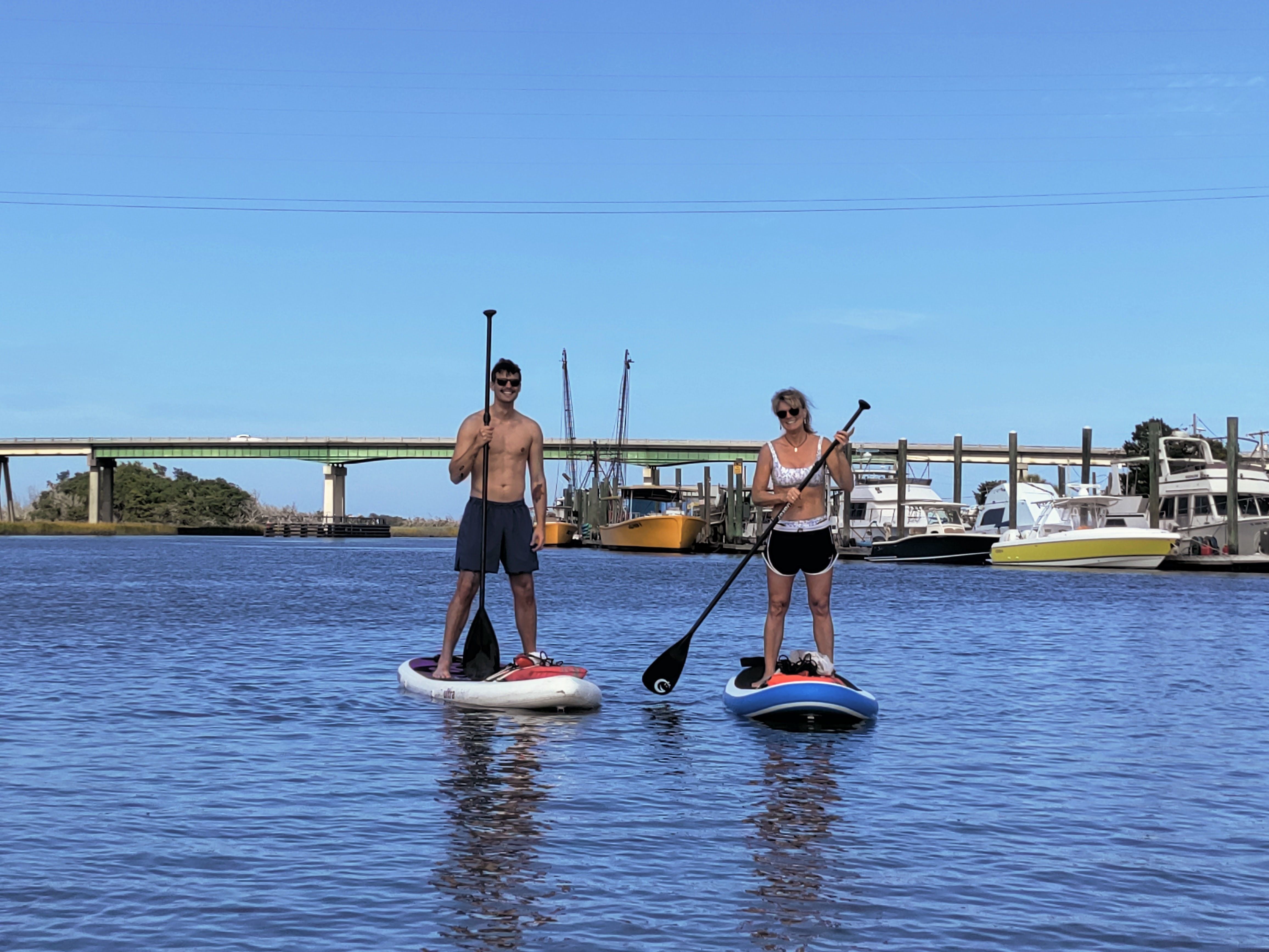 Two paddleboarders on stand-up boards in calm harbor water with docked boats and a low concrete bridge under a bright blue sky