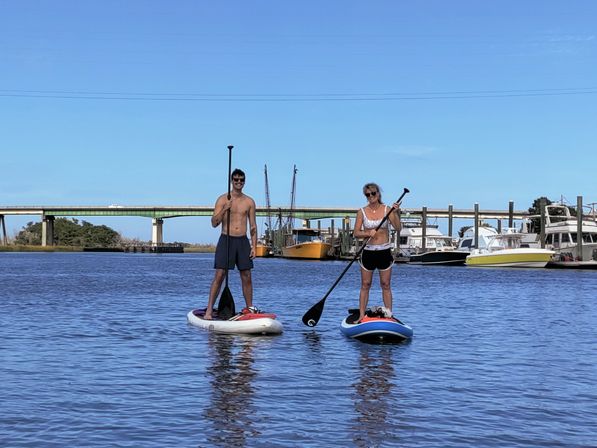 Two paddleboarders on stand-up boards in calm harbor water with docked boats and a low concrete bridge under a bright blue sky