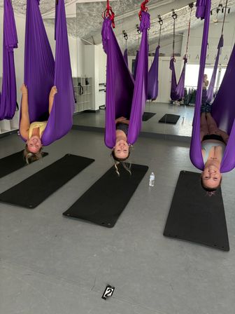 Three people smiling while hanging upside down in purple aerial yoga hammocks over black mats in a bright studio with a mirrored wall, aerial yoga class
