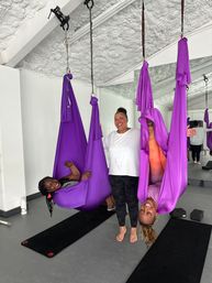 Three women smiling in an aerial yoga studio group class with bright purple silk hammocks; two participants suspended (one inverted) above black mats in front of a mirrored wall.