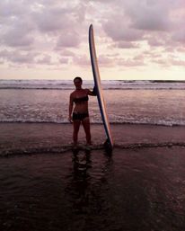 Surfer in a black bikini standing in shallow surf holding a longboard upright on a pink‑hued sunset beach with gentle ocean waves and a cloudy sky