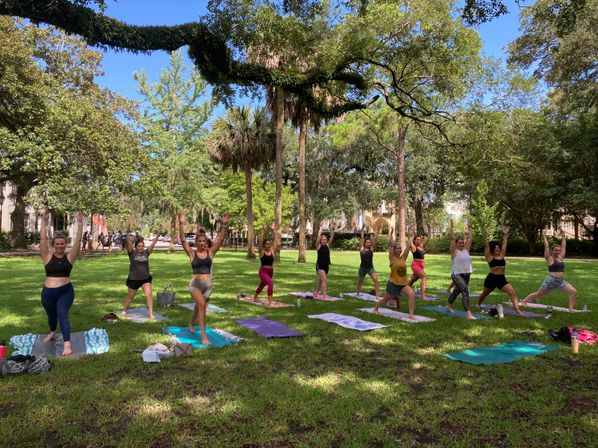 Outdoor yoga class in a sunny urban park: a group of people on colorful mats practicing warrior pose beneath live oak and palm trees on a lush green lawn.
