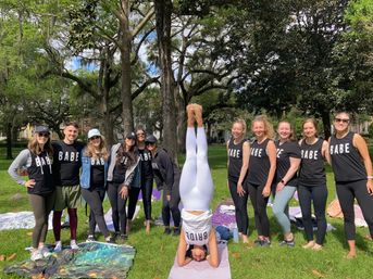 Bachelorette group photo in a shaded park — bride performing a headstand on a yoga mat while friends in matching “BABE” shirts pose around her under large oak trees with blankets and picnic gear.