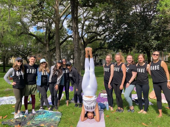 Bachelorette group photo in a shaded park — bride performing a headstand on a yoga mat while friends in matching “BABE” shirts pose around her under large oak trees with blankets and picnic gear.