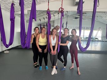 Six women in activewear smiling and posing among purple aerial yoga hammocks in a bright studio with a mirrored wall and gray floor — group aerial yoga class scene.