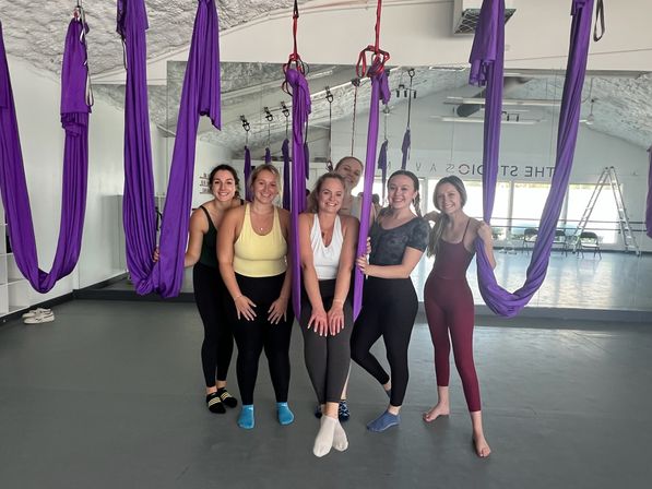 Six women in activewear smiling and posing among purple aerial yoga hammocks in a bright studio with a mirrored wall and gray floor — group aerial yoga class scene.