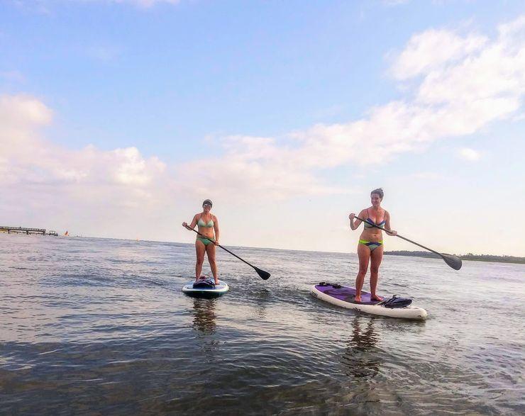 Two people stand-up paddleboarding on calm coastal waters under a bright blue sky with light clouds, shoreline visible in the distance.