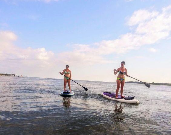 Two people stand-up paddleboarding on calm coastal waters under a bright blue sky with light clouds, shoreline visible in the distance.