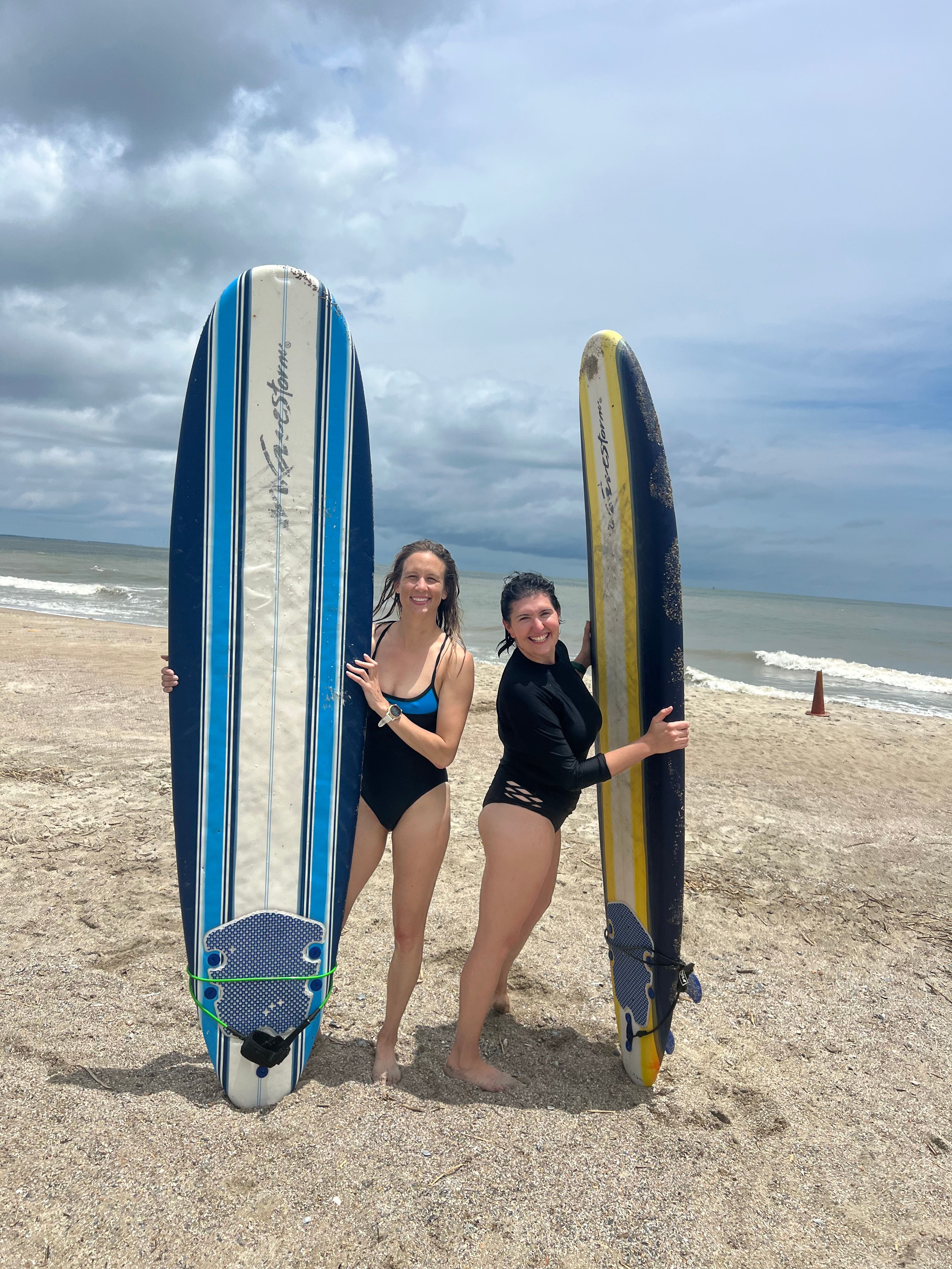 Two smiling women in swimsuits, barefoot and holding a blue-striped surfboard and a yellow surfboard, standing on a sandy beach with waves and a cloudy sky, ready to surf.