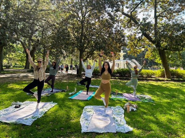 Five people practicing yoga in tree pose on mats on a sunny green lawn beneath large oak trees, with a decorative fountain and walking path in the background — outdoor group yoga class in a city park.
