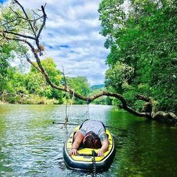 Relaxed paddler face-down on a yellow stand-up paddleboard in a calm river beneath an arching overhanging tree with a rope swing, surrounded by lush green forest and a cloudy blue sky.