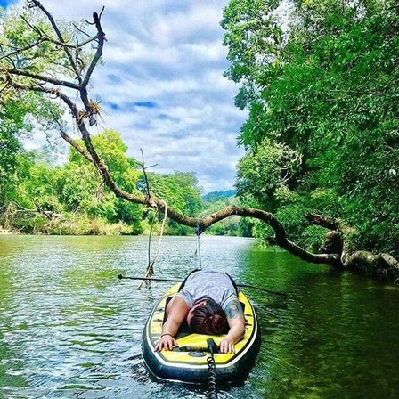 Relaxed paddler face-down on a yellow stand-up paddleboard in a calm river beneath an arching overhanging tree with a rope swing, surrounded by lush green forest and a cloudy blue sky.