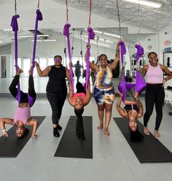 Group aerial yoga class in a bright mirrored fitness studio: six women in colorful activewear—three playfully suspended upside-down in purple silk hammocks above black mats while three stand and hold the silks.