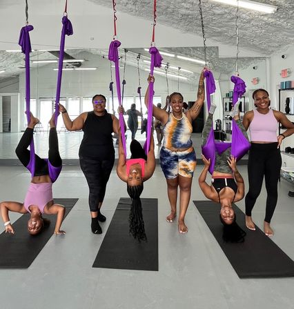 Group aerial yoga class in a bright mirrored fitness studio: six women in colorful activewear—three playfully suspended upside-down in purple silk hammocks above black mats while three stand and hold the silks.