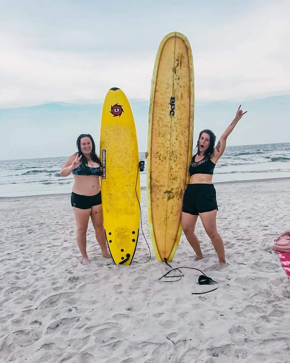 Two women in swimsuits on a sandy beach holding tall yellow surfboards with ocean waves behind them, one flashing a peace sign and the other cheering.