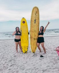 Two women in swimsuits on a sandy beach holding tall yellow surfboards with ocean waves behind them, one flashing a peace sign and the other cheering.