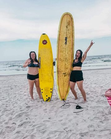 Two women in swimsuits on a sandy beach holding tall yellow surfboards with ocean waves behind them, one flashing a peace sign and the other cheering.