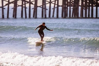 Surfer in a black wetsuit gliding on a longboard across a small wave with a wooden ocean pier in the background at a sunny beach