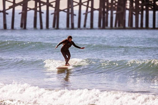 Surfer in a black wetsuit gliding on a longboard across a small wave with a wooden ocean pier in the background at a sunny beach