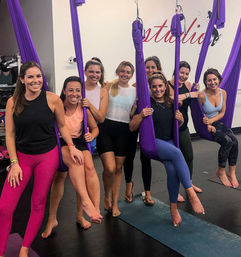 Smiling group of women in an indoor aerial yoga class using purple silk hammocks and yoga mats, wearing colorful activewear — group fitness studio scene.