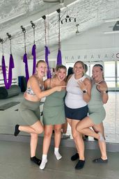Four smiling women in workout clothes pose and cheer inside an aerial yoga studio with purple silk hammocks hanging from the ceiling, one showing a ring while friends point, mirrored wall and fitness mats in the background