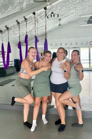 Four smiling women in workout clothes pose and cheer inside an aerial yoga studio with purple silk hammocks hanging from the ceiling, one showing a ring while friends point, mirrored wall and fitness mats in the background