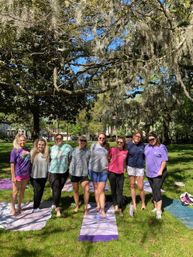 Nine women in activewear posing barefoot on yoga mats in a sunny oak‑shaded park draped with Spanish moss