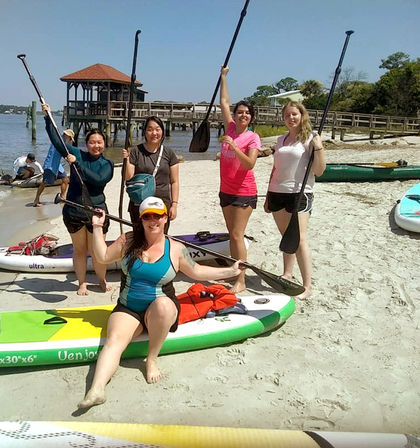 Group of five paddleboarders posing with paddles on a sunny sandy beach by a wooden pier, with stand-up paddleboards and kayaks on the shore and calm coastal water under a clear blue sky.