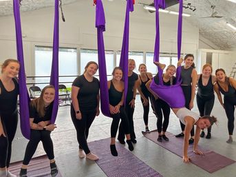 Aerial yoga group in a bright fitness studio: smiling women using purple aerial silks/hammocks over yoga mats, posing together with one student suspended upside down for a fun group workout.