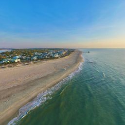 Aerial drone view of a quiet coastal town and long sandy beach curving toward a distant pier at sunset, gentle ocean waves lapping the shore.