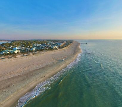 Aerial drone view of a quiet coastal town and long sandy beach curving toward a distant pier at sunset, gentle ocean waves lapping the shore.