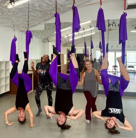 Three people inverted in purple aerial yoga hammocks in a bright studio with two smiling instructors beside them, mirrors and ceiling rigging visible — group aerial fitness class.