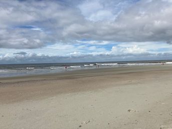 Wide sandy beach with gentle ocean waves under a dramatic cloudy sky, a few people walking along the shoreline and footprints in the foreground.