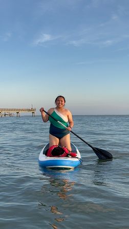Person smiling while standing on a blue stand-up paddleboard paddling on calm coastal waters near a wooden pier under a clear blue sky