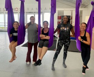 Five women smiling in a bright indoor aerial yoga studio in Savannah, posing in purple aerial hammocks and wearing activewear for a fun fitness class.