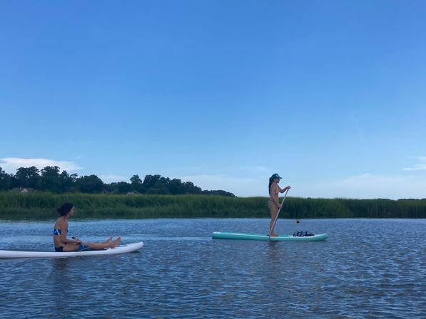 Two paddlers on stand-up paddleboards gliding through a calm coastal marsh with grassy shoreline under a bright blue summer sky