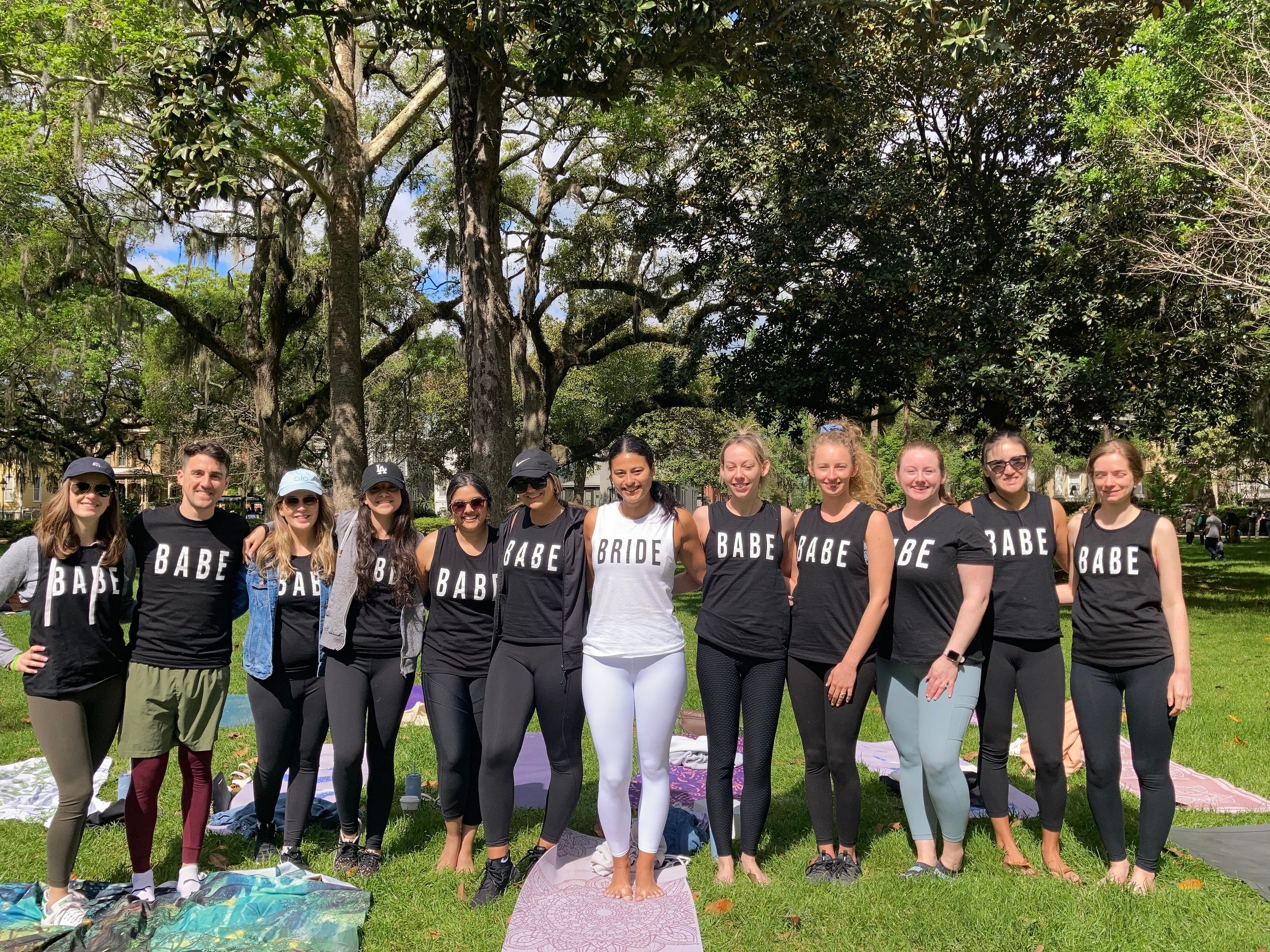 Bachelorette group wearing matching 'BABE' tanks with the bride-to-be in white, posing barefoot on yoga mats under large oak trees in a sunny park